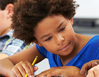 Boy holding pencil looks intently on as teaching assists with work at desk