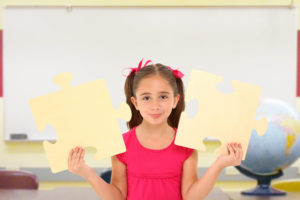Beautiful 7 year old girl holding two large puzzle pieces in school classroom.